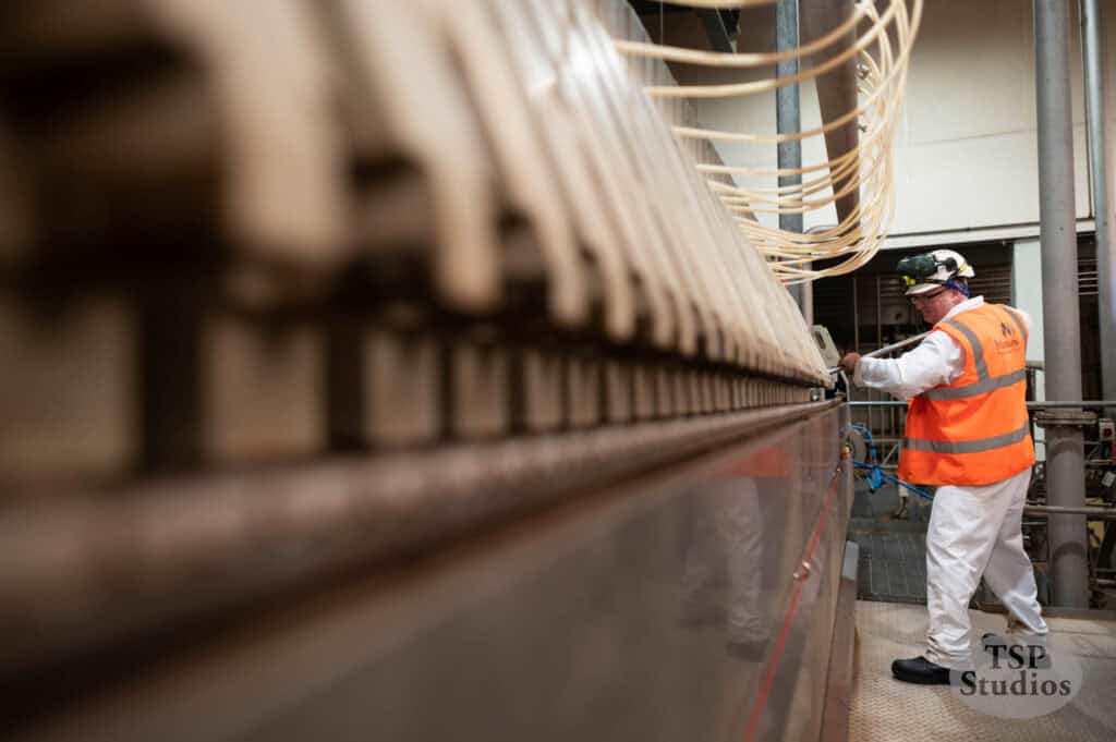 A worker in a white protective suit and orange safety vest operates equipment in an industrial setting. The worker is wearing a hard hat with an attached flashlight and safety glasses. Numerous cables and machinery components are visible in the foreground.