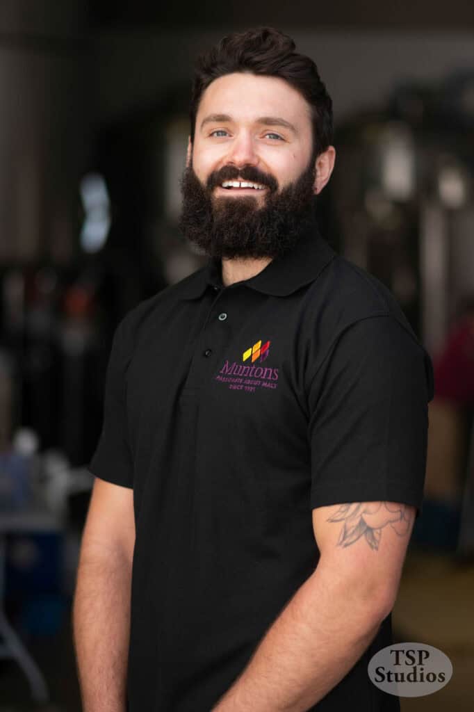 A man with a beard and mustache, wearing a black polo shirt with a Muntons logo on the left side of the chest, stands smiling. He has a tattoo on his left arm. The background is blurred, and the TSP Studios logo is in the bottom right corner of the image.