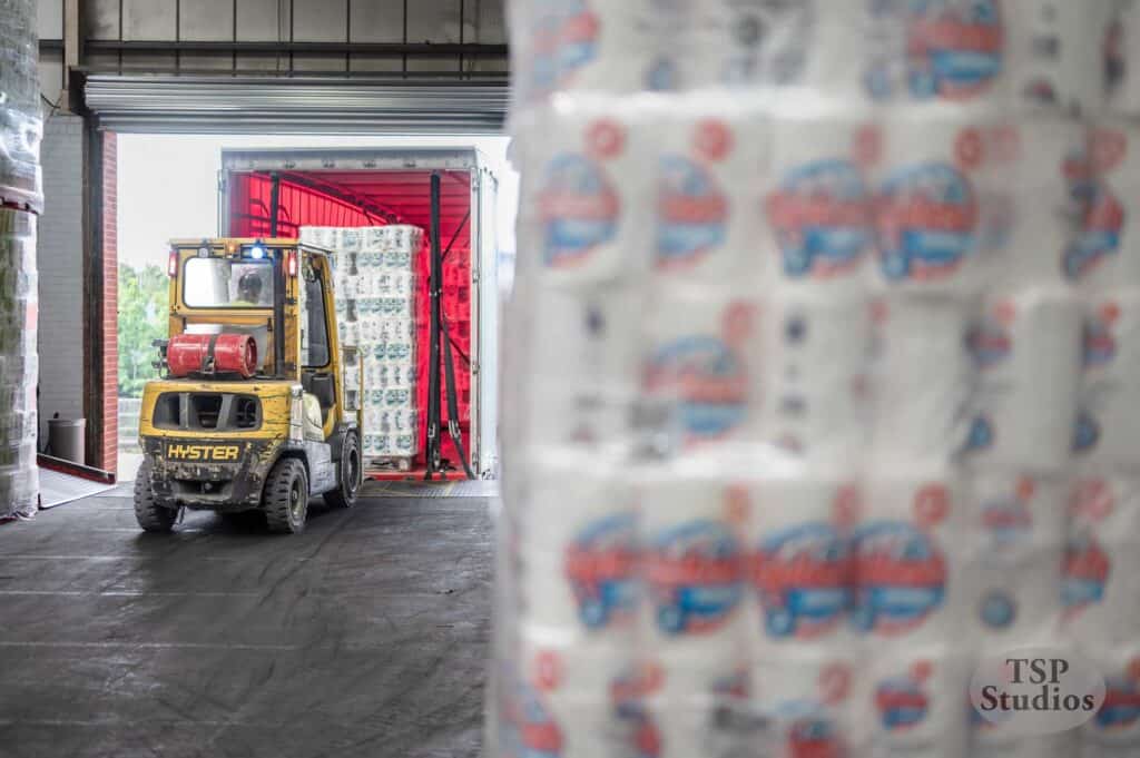 A forklift operator moves a pallet of packaged paper products into the back of a red truck inside a warehouse. In the foreground, there are stacks of similar packaged goods. Sunlight streams in from an open door, illuminating the scene.