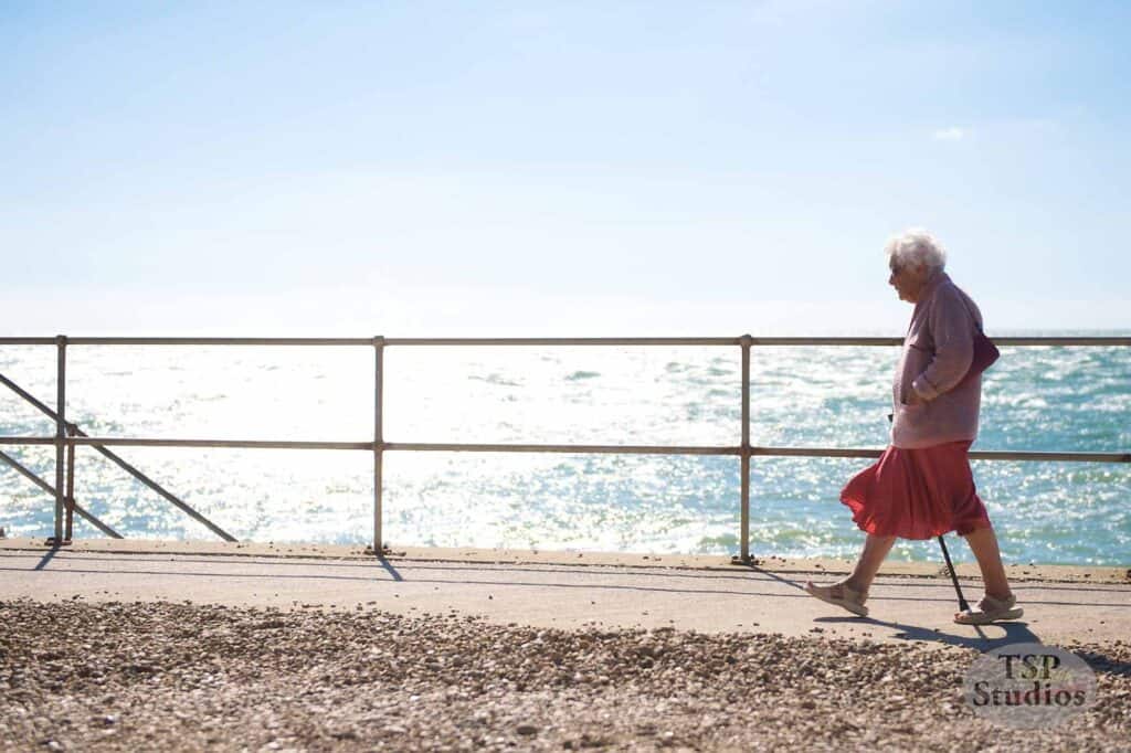 An elderly woman with white hair walks along a seaside promenade, using a cane for support. She wears a red skirt and pink cardigan. The bright sun shines on the sparkling sea in the background, creating a serene and peaceful atmosphere.