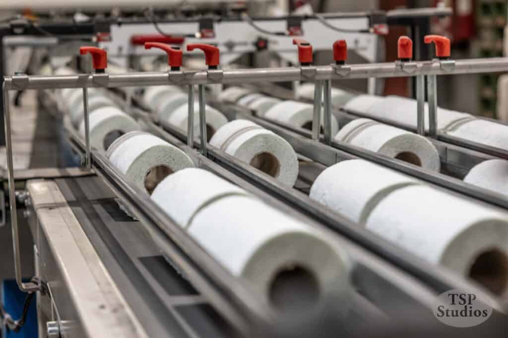 A close-up shot of a paper towel or toilet paper manufacturing line. Multiple paper rolls are aligned in rows on a conveyor belt system. Red clamps and metal bars are visible, holding and guiding the rolls through the machinery. The image has a TSP Studios watermark.