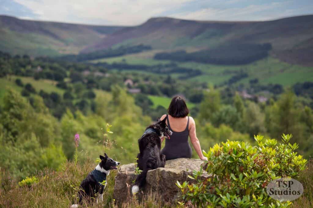 A person with short dark hair sits on a rock overlooking a lush, hilly landscape with two black and white dogs, one sitting next to them and the other standing nearby. The scene is serene, with vibrant greenery and a small valley visible in the distance.