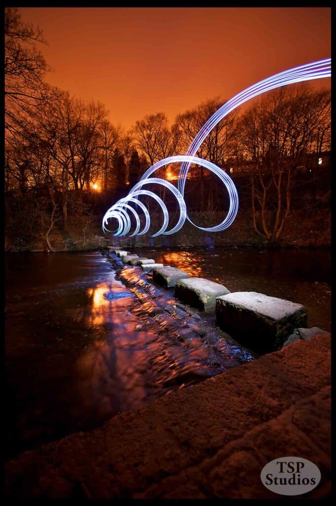 A striking long-exposure nighttime photograph shows a spiral of bright white lights hovering over a serene river with stepping stones. The sky glows with a warm orange hue, and dark silhouettes of trees frame the background. "TSP Studios" is branded in the lower right corner.