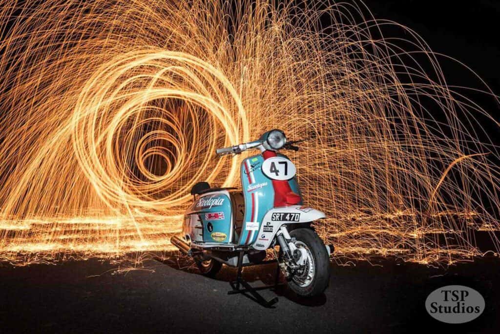 A vintage scooter with a racing number 47 and several stickers is parked on a dark background. Behind it, a spectacular long-exposure light effect creates swirling circles of bright orange light, adding dynamic motion to the scene. The image is credited to TSP Studios.