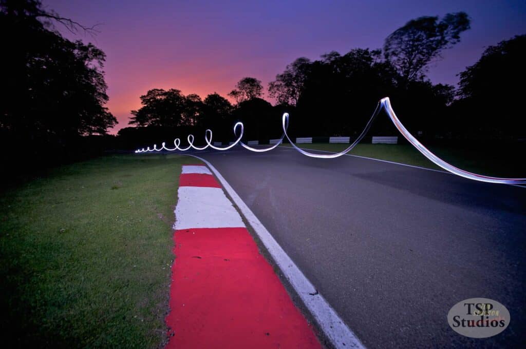 A stunning long-exposure photo captures a mesmerizing light trail twisting above a race track as twilight sets in, with a purple and orange sky. The track curves to the right, lined with red and white curbs, and framed by dark silhouettes of trees.