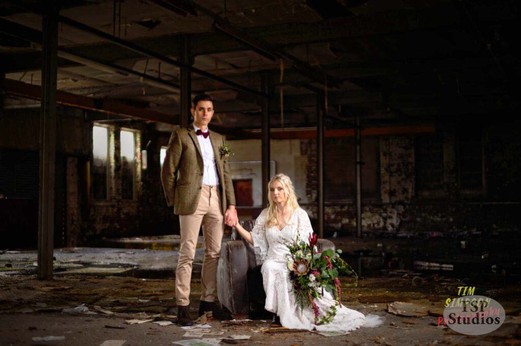 A man in a suit and a woman in a white dress, holding a large bouquet of flowers, pose in an abandoned, dilapidated building. The man stands with one hand on a suitcase, while the woman sits beside it on the ground, surrounded by scattered debris.