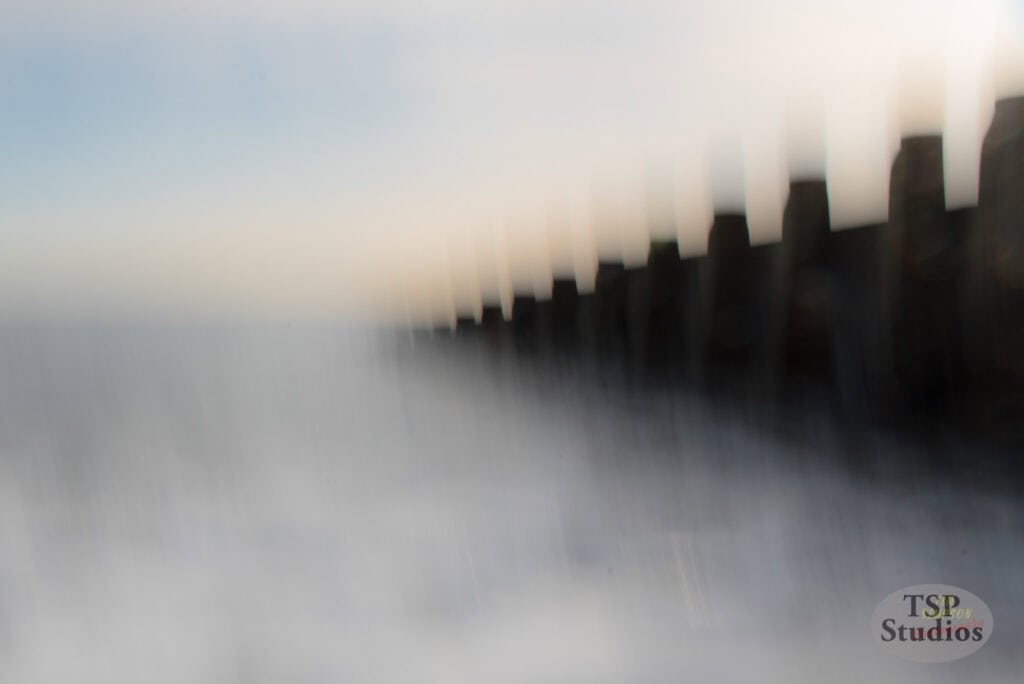 A long-exposure photo showing a row of dark vertical structures gradually fading into the distance, against a backdrop of blurred white and blue hues, giving a dreamy, ethereal effect. The image has the text "TSP Studios" in the bottom right corner.