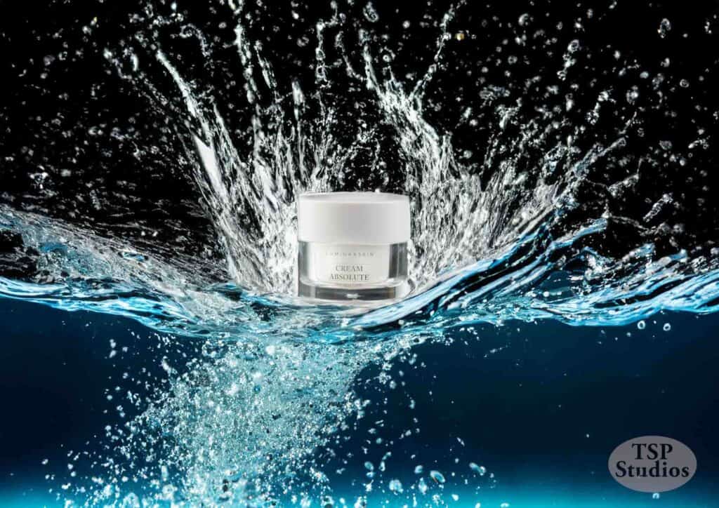 A white jar of face cream is captured mid-splash in crystal-clear water against a black background, creating an energetic explosion of droplets around it. The text "TSP Studios" is visible in the lower right corner.