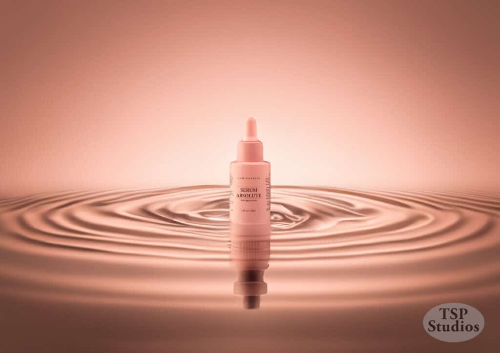 A sleek, pink skincare serum bottle stands upright in the center of a rippling pool of liquid. The bottle features a dropper cap, and the background is a warm, soft gradient of pink hues. The label on the bottle reads "Serum Absolut." TSP Studios logo is seen.