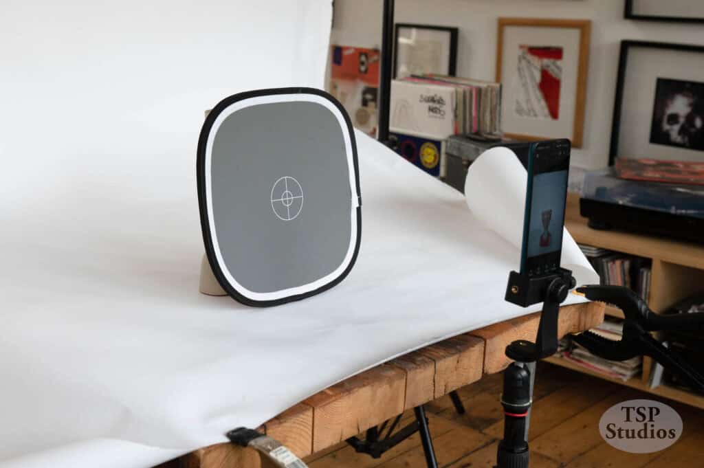 A photography setup with a gray calibration card on a table, surrounded by white backdrop material. A smartphone on a tripod is positioned to capture the card. The background has framed artworks and shelves with various items. The scene appears to be in a studio.