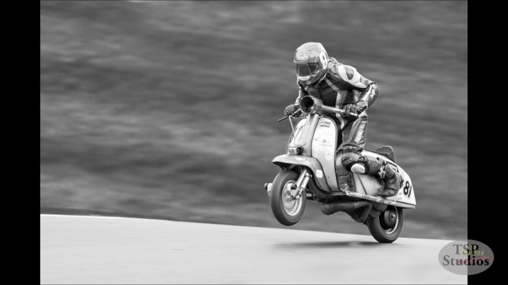 A black and white photo of a motorcyclist performing a jump on a vintage scooter. The rider, dressed in racing gear and helmet, is airborne with the scooter tilted slightly forward, against a blurred background. The logo "TSP Studios" is visible in the bottom right corner.