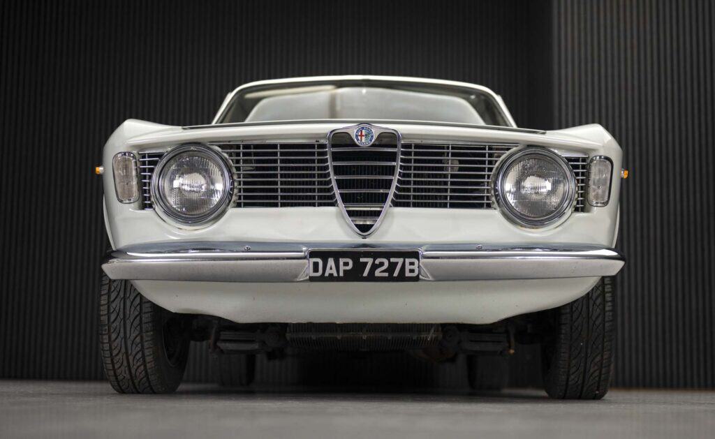 A classic white car viewed from the front, displaying a vintage design with round headlights and a distinctive grille. The license plate reads "DAP 727B." The background features dark vertical lines.