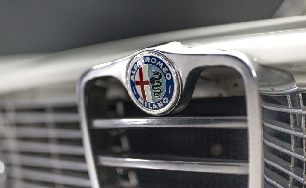 Close-up of an Alfa Romeo emblem on the front grille of a white car. The badge features the iconic red cross and serpent design with the words "ALFA ROMEO MILANO" encircling it. The shiny metallic grille is prominently displayed.