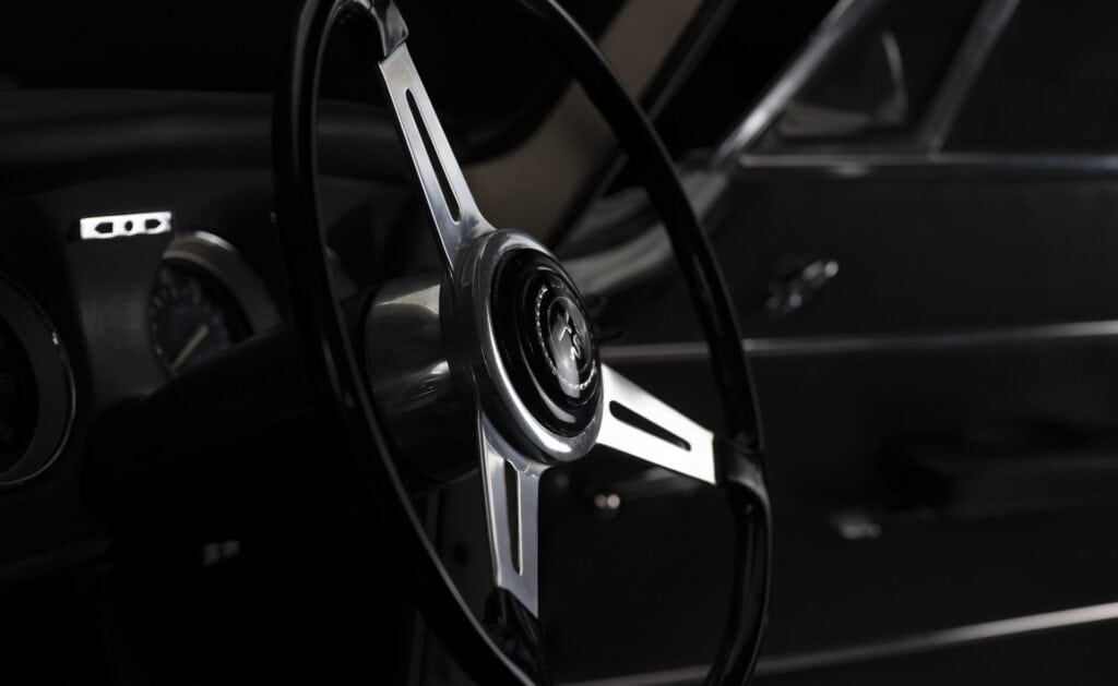 A close-up of a classic car's steering wheel, featuring a shiny chrome center with a horn button. The dashboard and part of the car door are visible in the dimly lit interior, creating a vintage atmosphere.